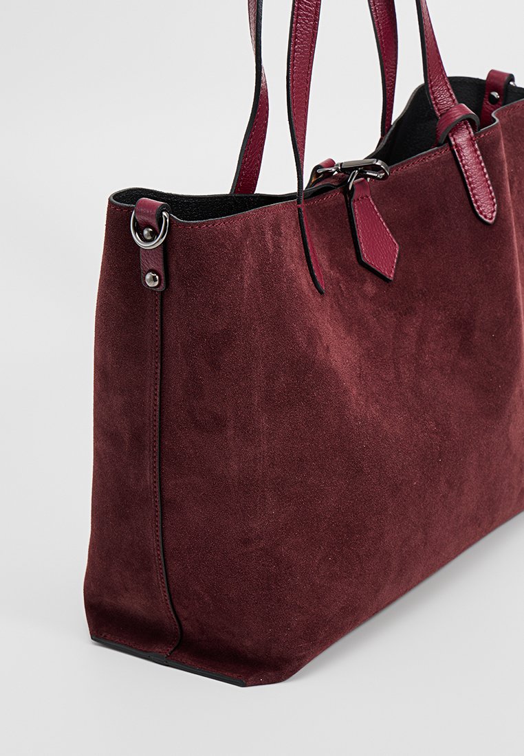 Bordeauxfarbene Wildleder-Tasche mit Lederelementen, doppelten Schulterriemen und silbernem Beschlag. Verfügt über eine sanfte Textur und eine strukturierte Form.