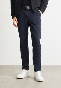 Navy blue cotton trousers with a slim fit, worn with a black belt and white sneakers. Smooth texture, tailored design, and minimal detailing.