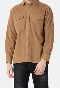Bruine corduroy shirt met lange mouwen, knoopsluiting aan de voorkant, twee borstzakken en een subtiele ribbelstructuur. Knopen in een contrasterende kleur.