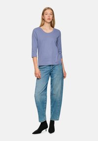 Helllila, langärmliges Oberteil mit Rundhalsausschnitt, kombiniert mit lockeren blauen Jeans. Das Outfit wird mit schwarzen Knöchelstiefeln abgerundet.