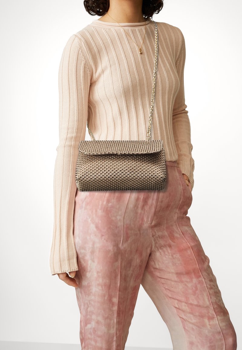 Personne portant un haut beige côtelé à manches longues, un pantalon rose à motifs, un collier en or et portant un sac à épaule beige tissé avec une bandoulière chaîne.