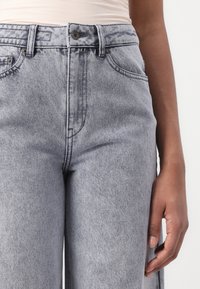 Jeans a vita alta di colore grigio chiaro con design a gamba dritta. Include chiusura con zip, bottone e tasche anteriori e posteriori.