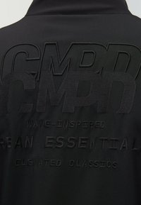 Black jacket featuring embossed designs with the text "CMPD" and "WAVE-INSPIRED URBAN ESSENTIALS." Smooth texture and simple silhouette.