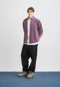 Fialová kordové košeľa, pod ňou biela tričko, čierne voľné nohavice, béžové topánky; stojí na svetlom pozadí s modrou podlahou.