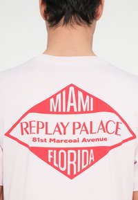 Πίσω μέρος λευκού μπλουζομένάς με κόκκινο σχέδιο σε σχήμα ρόμβου που αναγράφει "Miami Replay Palace 81st Marcoai Avenue Florida."