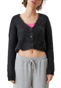 Černý zkrácený cardigan s chlupatou strukturou, véčkovým výstřihem a třemi knoflíky, nošený přes růžový top a šedé tepláky.