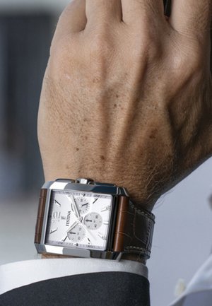 Montre carrée en argent avec cadran blanc, bracelet en cuir marron, fonctions chronographe et accents en métal poli, portée au poignet.