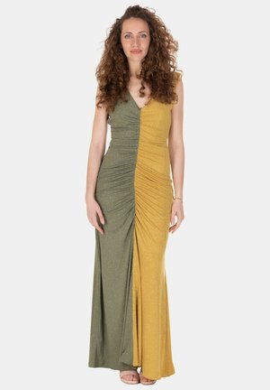 Donna con capelli ricci che indossa un abito senza maniche lungo fino a terra, bicolore con pannelli verde oliva e giallo senape, in piedi davanti a uno sfondo semplice.