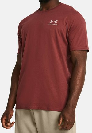 Mann trägt ein bordeauxfarbenes kurzärmeliges Under Armour Rundhals-Shirt und hellbeige Hose vor einfachem Hintergrund.
