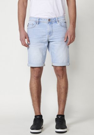 Pantalones cortos de mezclilla de color azul claro con dobladillos enrollados, cinco bolsillos, cierre frontal de botón y un ligero desgaste en la tela. Zapatillas negras puestas.