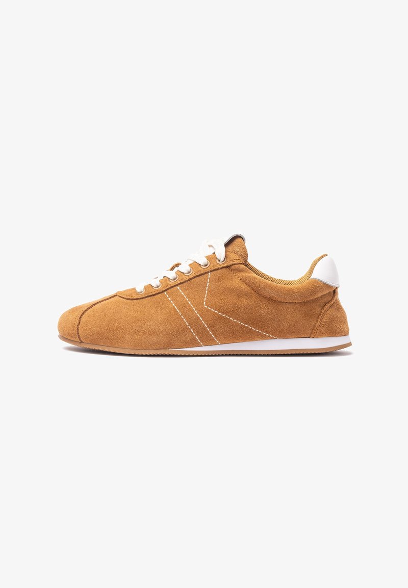 Brown suede sneaker with white lace-ups, contrasting white stitching, and a white heel accent. Low-top design with a flexible sole.