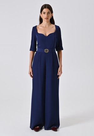 Marineblauer maßgeschneiderter Jumpsuit mit Herzausschnitt, kurzen Ärmeln und einem breiten Gürtel mit einer runden goldenen Schnalle. Glattes, strukturiertes Material.