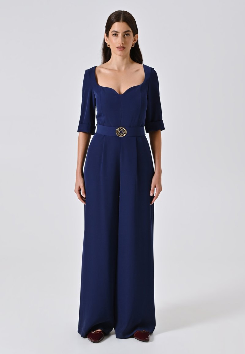 Navyblauwe op maat gemaakte jumpsuit met een sweetheart-uitgesneden halslijn, korte mouwen en een brede riem met een ronde gouden gespsluiting. Gladde, gestructureerde stof.