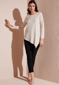 Top a maniche lunghe color crema con orlo asimmetrico, abbinato a pantaloni neri slim fit e tacchi a fascia, davanti a una parete in pesca testurizzata.