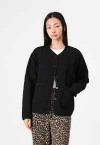 Black quilted jacket with a round neckline, button front, and two patch pockets. Worn over a white top and patterned leopard-print pants.