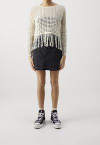 Szydełkowy kremowy sweter z frędzlami na dole, w połączeniu z dopasowaną czarną spódnicą dżinsową i czarnymi butami sportowymi za kostkę z białą podeszwą.