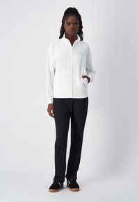 Giacca bianca con zip e colletto alto, abbinata a pantaloni neri a gamba dritta e sneakers nere. Tessuto liscio, design minimale.