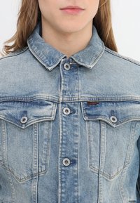 Ung person som bär en ljusblå denimjacka med knäppta krage och framfickor, lång brunt hår synligt.