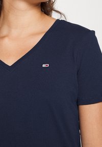 Námořnická modrá bavlněná tričko s výstřihem do V, krátkými rukávy a malou nášivkou s logem v červené, bílé a modré barvě na levé straně hrudi.