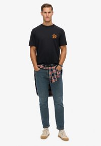 Camiseta de algodón negra con un logo naranja en el pecho, combinada con jeans de color azul claro y una camisa de cuadros atada a la cintura. Zapatillas beige.