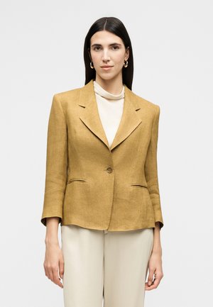 Ženska z dolgimi temnimi lasmi, ki nosi zlate obročne uhane, gorčico blazer, bež hlače z visokim pasom in svetel rolka, stoji pred enostavnim ozadjem.