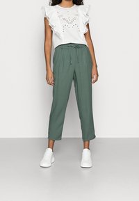 Blusa blanca con volantes y detalles de encaje, combinada con pantalones holgados de color verde olivo. La modelo lleva zapatillas blancas. Fondo minimalista.