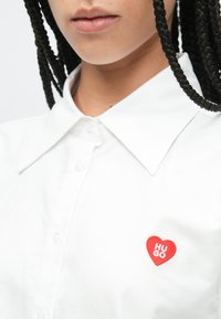 Bijela košulja s ovratnikom s crvenim zakrpanim srcem s tekstom "HUGO". Glatka tekstura tkanine s kopčama na gumbe.