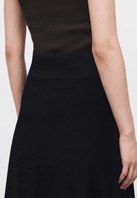 Femme portant un haut noir sans manches et une jupe noire taille haute, vue de dos, les bras détendus le long du corps.