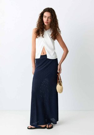 Donna con capelli ricci che indossa un top bianco senza maniche a fascia corta, gonna lunga blu navy, sandali neri e tiene una piccola borsa intrecciata.