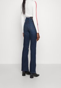 Henkilö, jolla on pitkät letit, pukeutuneena tummansinisiin korkeavyötäröisiin farkkuihin, mustiin korollisiin saappaisiin ja valkoiseen neulepuseroon, jossa on punaiset raidat hihoissa, seisoo.