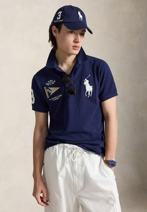 Νέος άνδρας που φοράει μπλε πόλο με μεγάλο λογότυπο άσπρου αλόγου, αντίστοιχο καπέλο, λευκό παντελόνι με κορδόνι, γυαλιά ηλίου και μπλε ρολόι χειρός, ποζάρει χαλαρά.