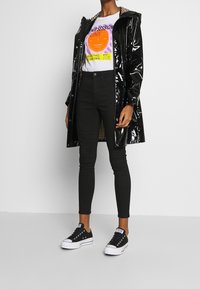 Person trägt schwarzen glänzenden Regenmantel über weißem T-Shirt mit Grafik, schwarze enge Jeans und schwarz-weiße Sneakers und steht auf einfachem Boden.