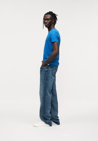 Μπλε βαμβακερό μπλουζάκι, χαλαρή εφαρμογή, συνδυασμένο με τζιν παντελόνι ανοιχτού γαλάζιου. Το μοντέλο φοράει άσπρα αθλητικά παπούτσια. Απλός σχεδιασμός, ουδέτερο φόντο.