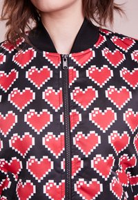 Bomber jacket featuring a black background with a pixelated red heart pattern. Zippered front with black ribbed collar and cuffs.