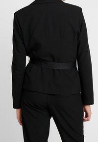 Blazer negro a medida con silueta ajustada, mangas largas y cinturón de satén en la cintura. Textura suave y diseño clásico de cuello.