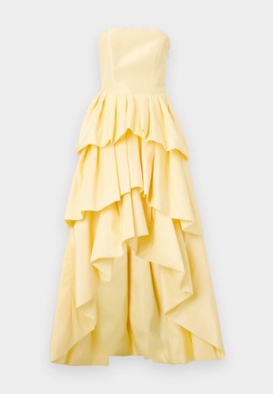 Bezplecu dzeltena kleita, kas izceļas ar slāņainiem volāniem, pieguļošu augšdaļu un rakstainu audumu. Volāni veido plūstošu siluetu apakšmalā.