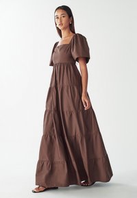 Brown maxi dress with puffed sleeves and a square neckline. Features tiered design and smooth texture. Worn with black sandals.