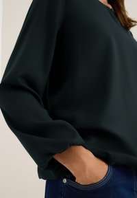 Donkergroene blouse met losse, wijd uitlopende mouwen en een ronde halslijn, gemaakt van een zachte stof; gelaagd over blauwe denim jeans.