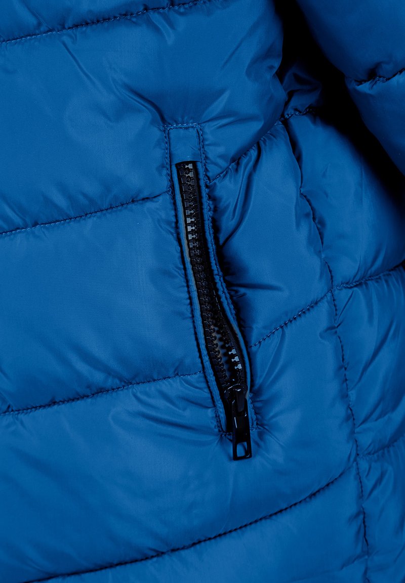 Veste rembourrée bleue avec une poche zippée noire. Le tissu a une texture lisse et comporte des sections matelassées pour l'isolation.