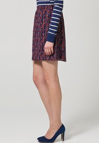 Jupe imprimée avec un fond bleu marine, ornée de motifs de pois rouge et blanc. Taille élastique, longueur au-dessus du genou, associée à des escarpins bleus.