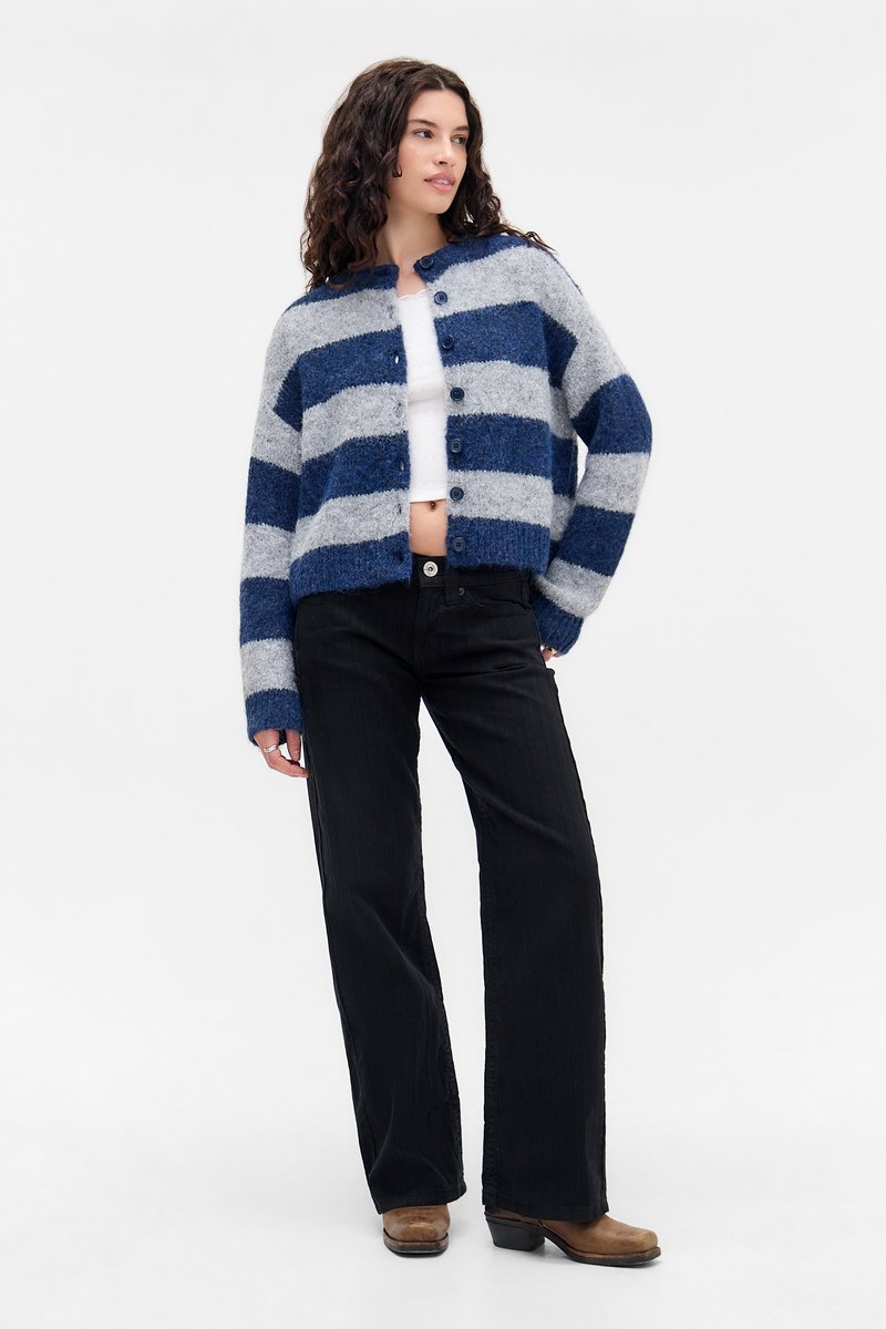 Vrouw met krullend haar, gekleed in een blauw en grijs gestreept knopentrui, witte cropped top, zwarte wijde spijkerbroek en bruine laarzen, staand.