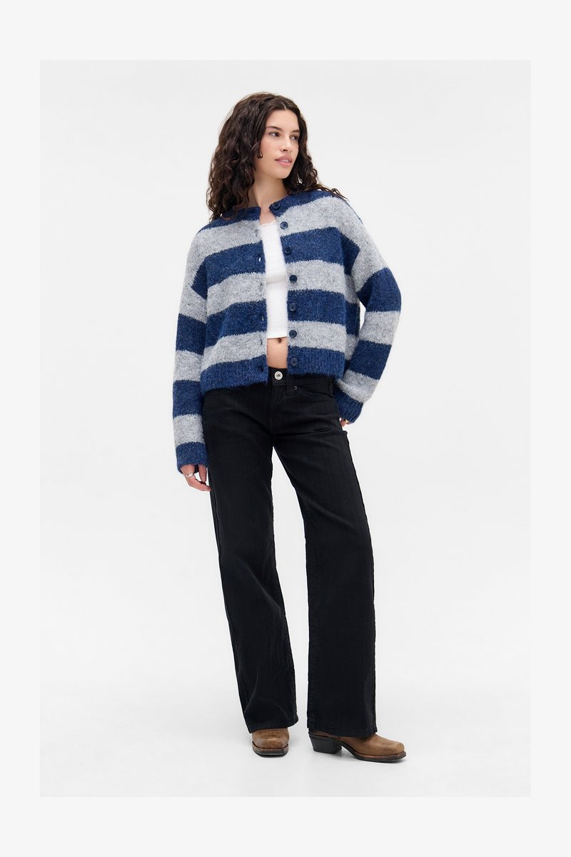 Kortsukirju ja halli triibulise nööbiga kampsuniga naine, valges lühikeses topsis, mustades laia säärega teksastes ja pruunides saapastes, seistes.