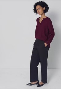 Vínově červený top s dlouhým rukávem a výstřihem do V, doplněný černými širokými kalhotami a černými špičatými balerínami, na neutrálním pozadí.