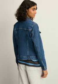 Denim jacket in dark blue, featuring a classic cut, long sleeves, and buttoned cuffs. The fabric has a textured finish with stitching details.