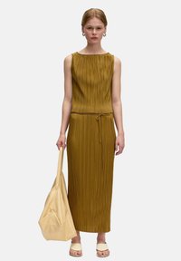Senffarbenes ärmelloses Faltenkleid mit einem Bindegürtel. Das Model trägt eine beigefarbene lässige Tasche und leichte Sandalen.