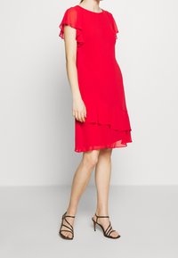 Femme portant une robe rouge jusqu'aux genoux avec des manches et un ourlet volantés, associée à des sandales à talons hauts noires à lanières, se tenant sur un fond uni.