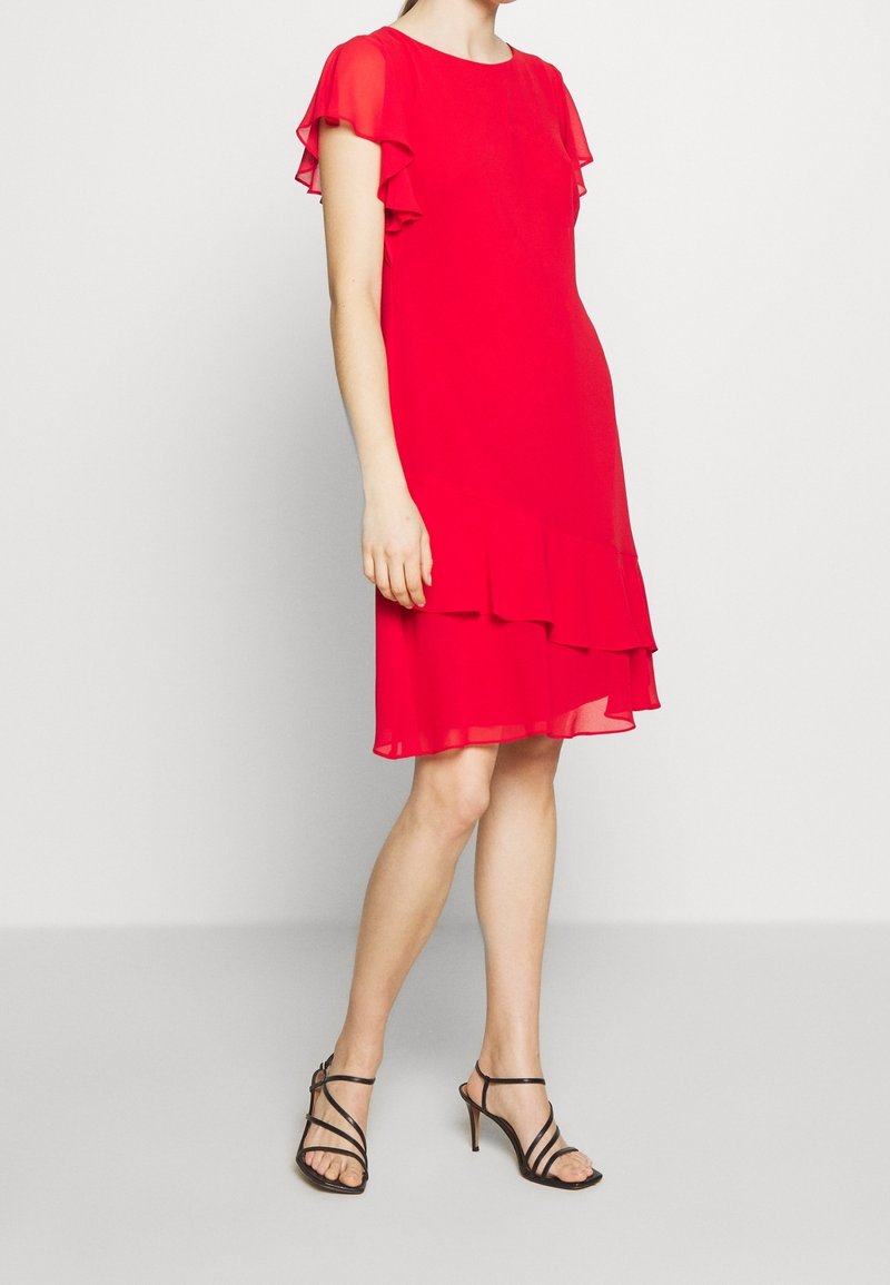 Femme portant une robe rouge jusqu'aux genoux avec des manches et un ourlet volantés, associée à des sandales à talons hauts noires à lanières, se tenant sur un fond uni.