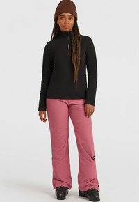 Woman wearing brown beanie, black zip-up sweater, pink snow pants, and black ski boots standing against plain white background.