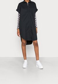 Abito camicia nero con bottoni frontali, maniche corte arrotolate, aperture laterali e una tasca applicata, indossato sopra una maglietta a maniche lunghe con motivo.