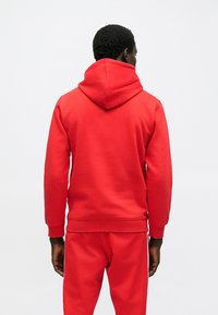 Muži s tmavou pokožkou, ktorý nosí červenú mikinu s kapucňou a zodpovedajúce červené nohavice, stojí chrbtom k nám na bielom pozadí.